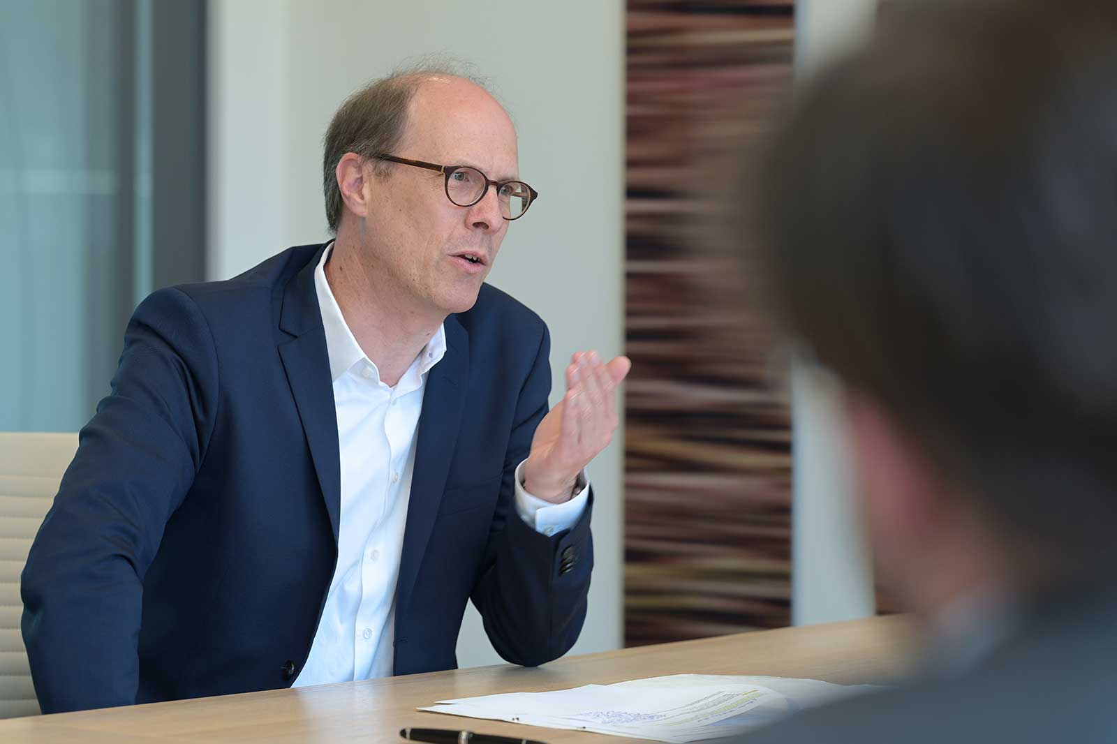 A man in a suit sits at a table, gesturing during a discussion in a modern office.
