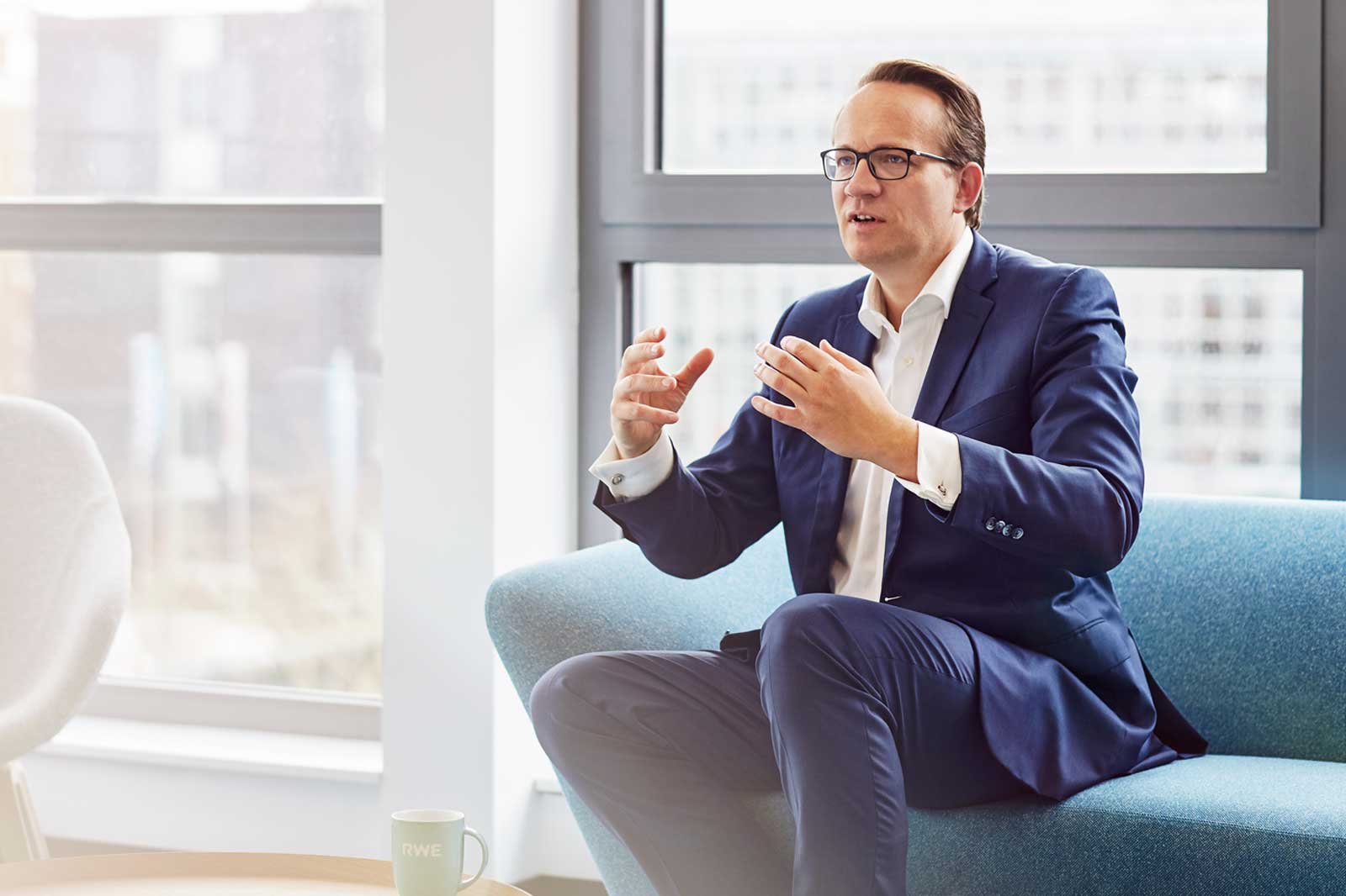 Ein Mann in einem Anzug gestikuliert, während er auf einem blauen Sofa in einem modernen Büro mit großen Fenstern sitzt.