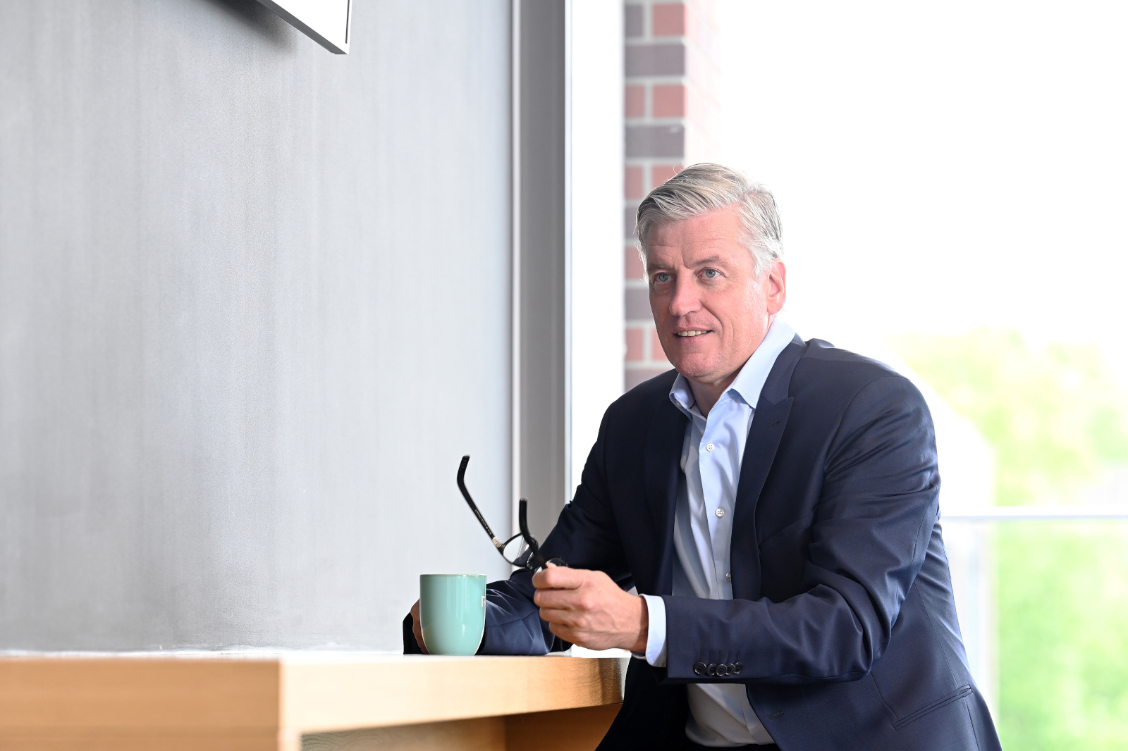 Ein Mann in einem Anzug sitzt an einem Tisch mit einer Tasse und einer Brille in der Hand.