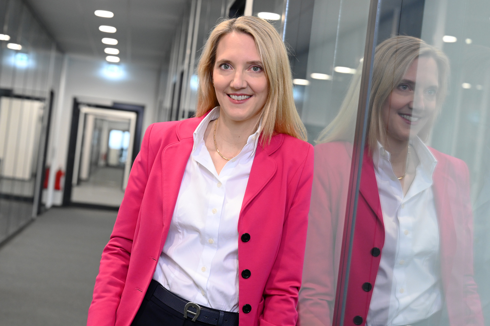 Eine Frau in einem pinken Blazer steht in einem modernen Büro mit Glaswänden und neutralem Teppich.