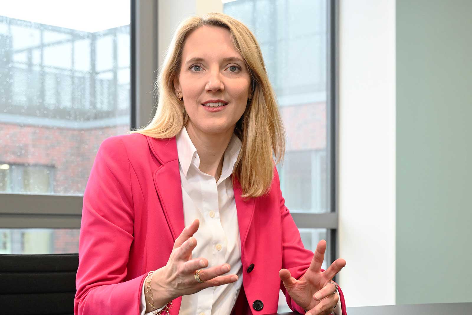 A person with long blonde hair is wearing a pink blazer and is gesturing while speaking in a modern office.