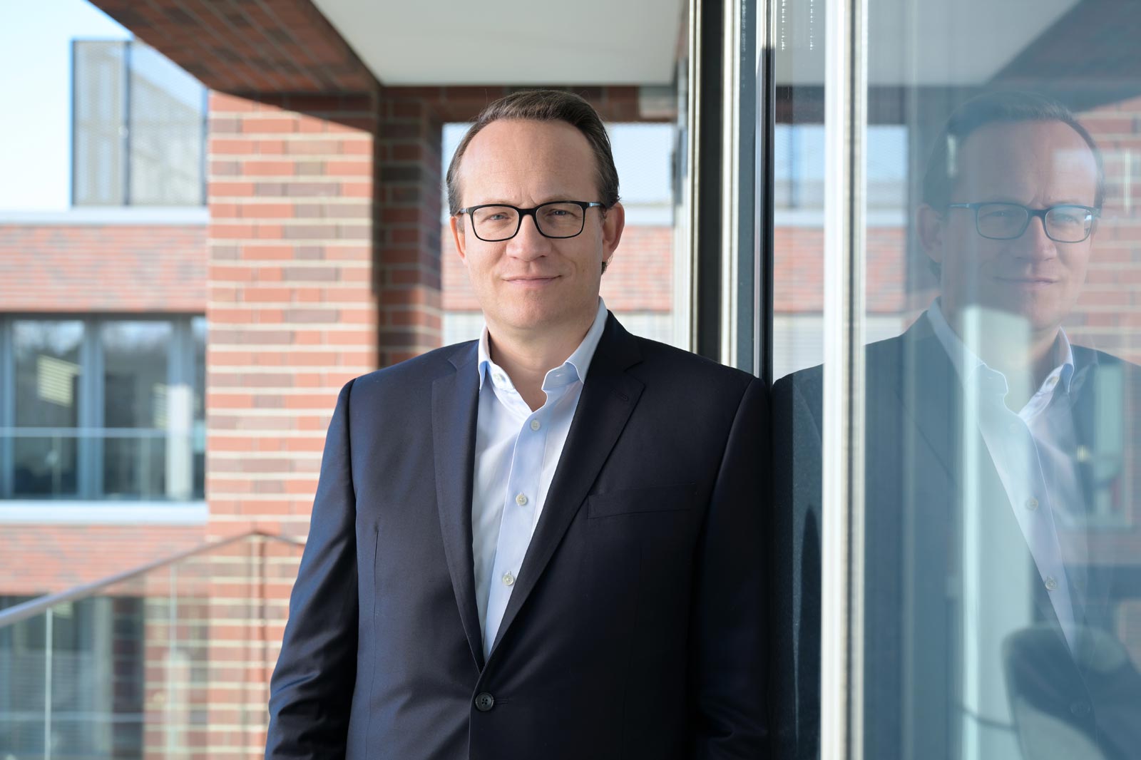 A man in a dark suit stands beside a glass wall in a modern building. Portrait of Markus Krebber.