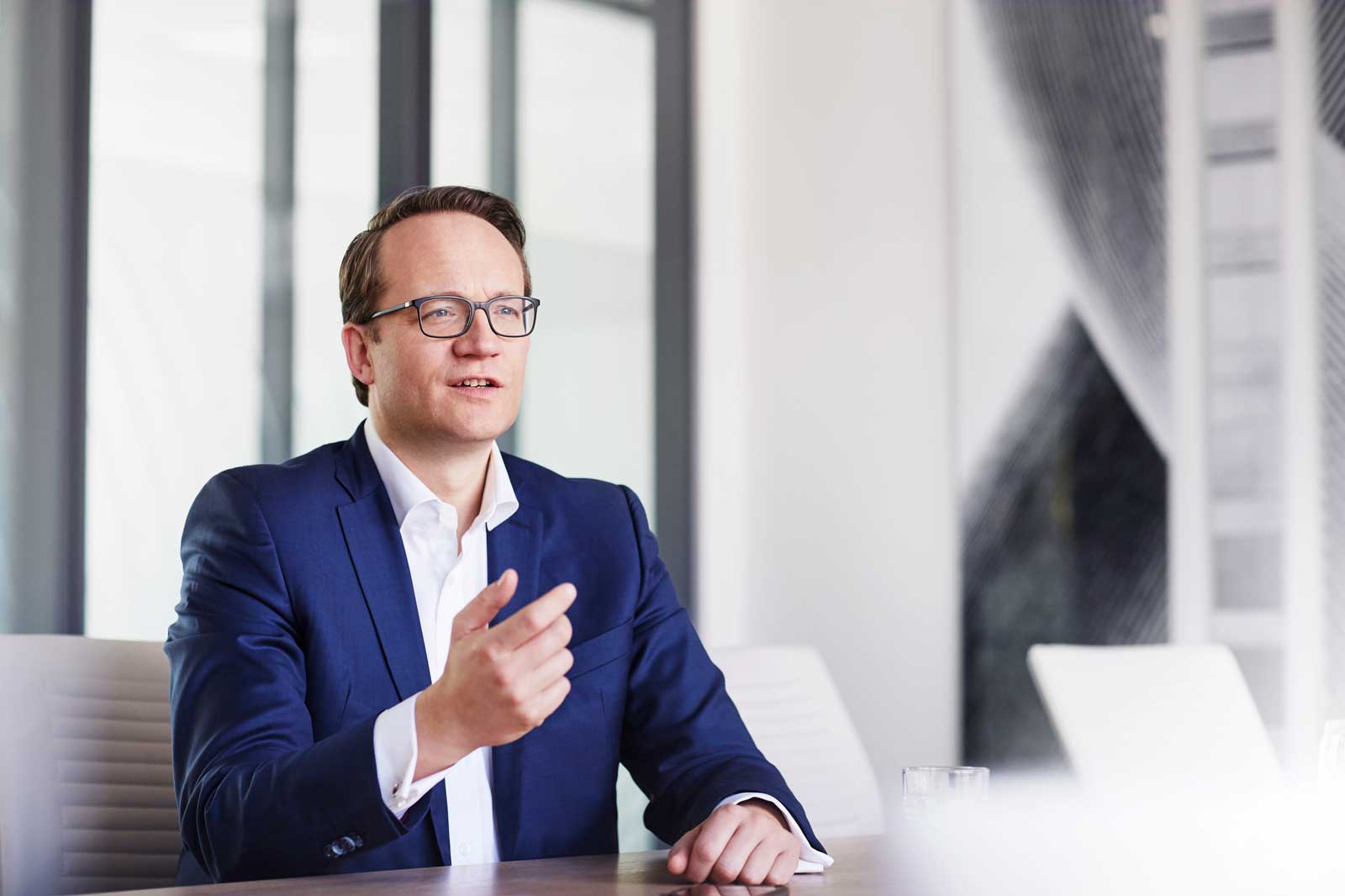 Ein Mann im blauen Anzug gestikuliert, während er an einem Konferenztisch in einem modernen Büro sitzt.