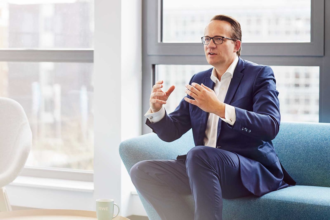 Ein Mann in einem Anzug gestikuliert, während er auf einem blauen Sofa in einem modernen Büro mit großen Fenstern sitzt.