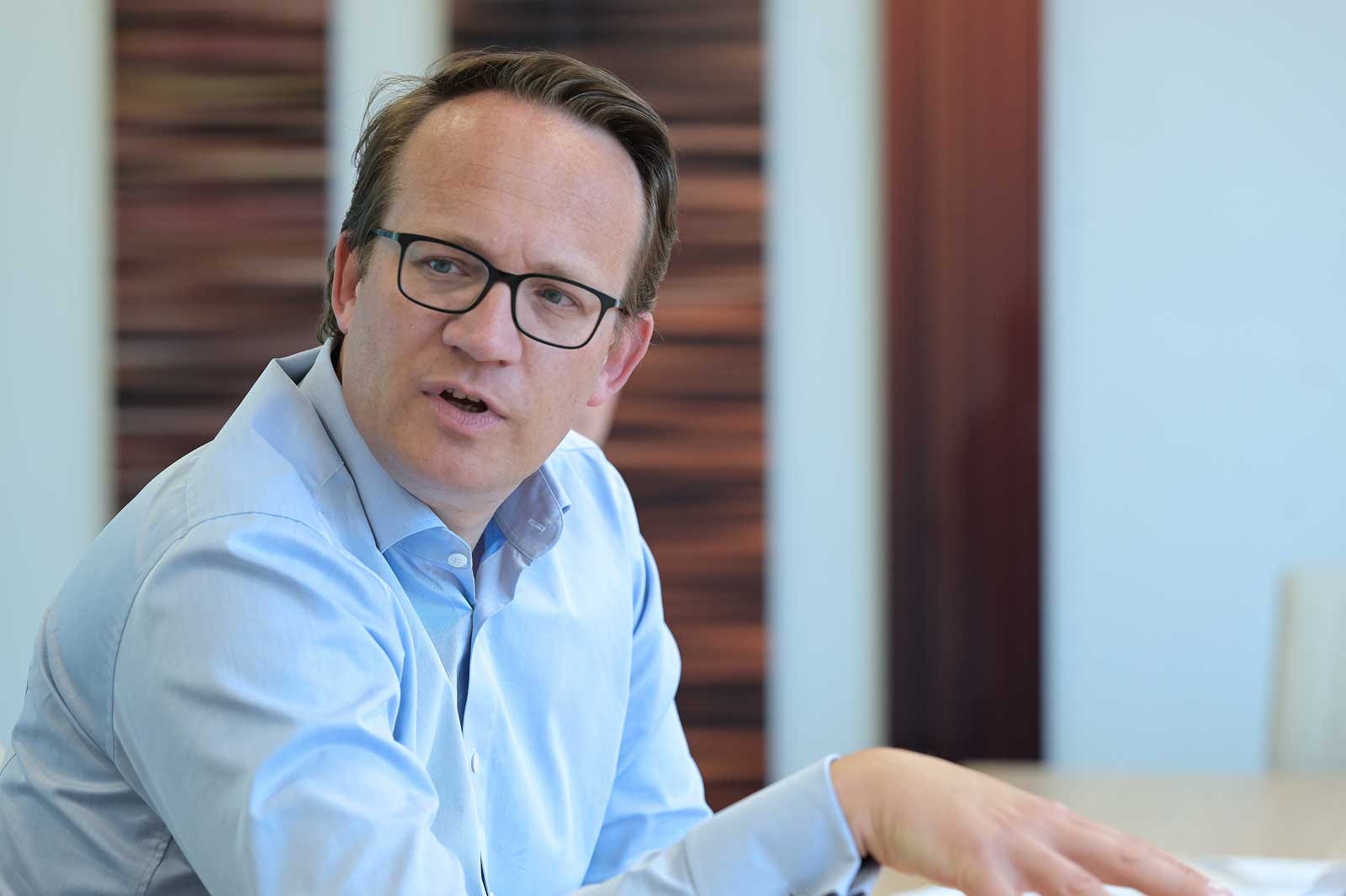 A man in a light blue shirt gestures while seated at a table in a modern office setting with wooden panels in the background.