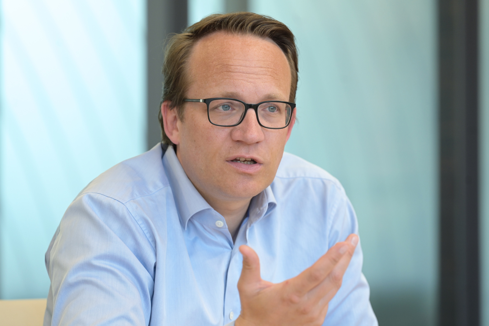 Ein Mann in einem hellblauen Hemd gestikuliert, während er an einem Konferenztisch in einem modernen Büro sitzt.