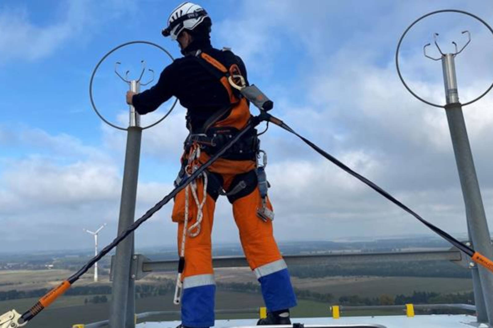 Ein Techniker in Sicherheitsausrüstung arbeitet in großer Höhe an Geräten und hat einen weiten Blick über die Landschaft.