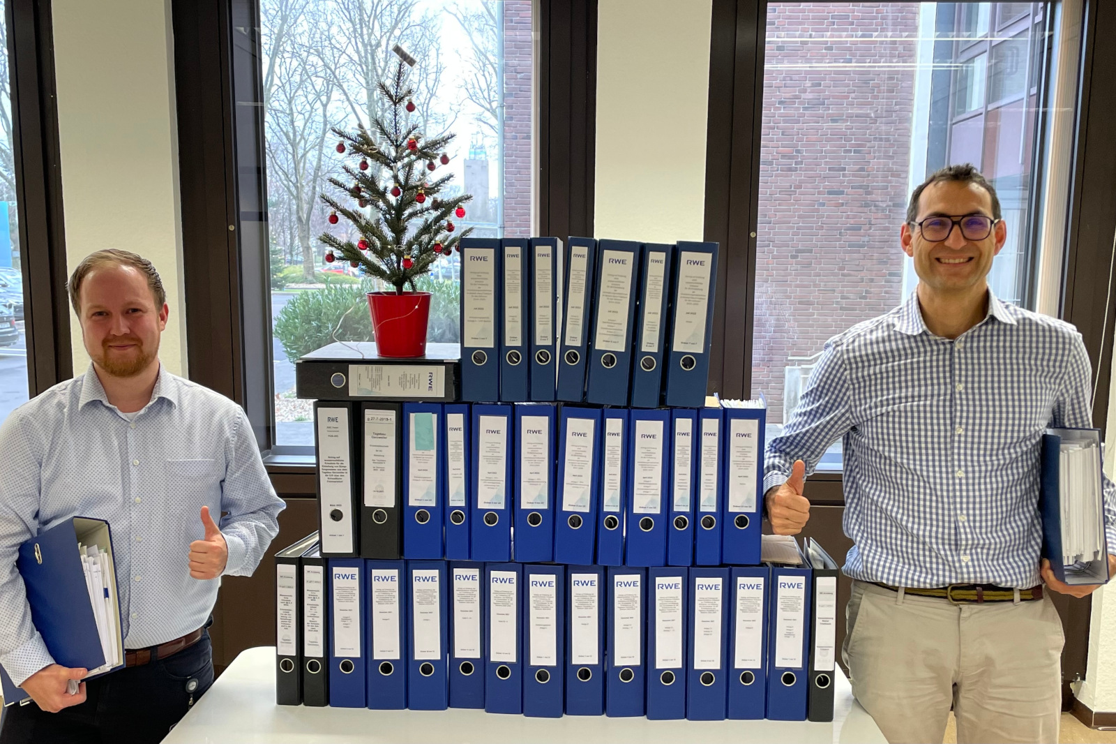 Zwei Personen stehen neben einem großen Stapel blauer Ordner und einem kleinen Weihnachtsbaum auf einem Tisch in einem Büroraum.