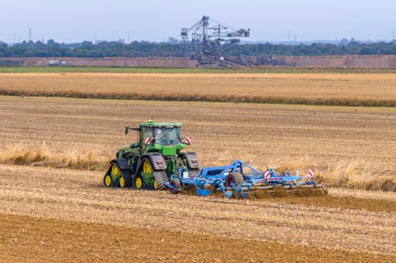 Ein grüner Traktor ploughed ein Feld, mit einer großen Industriemaschine im Hintergrund sichtbar.