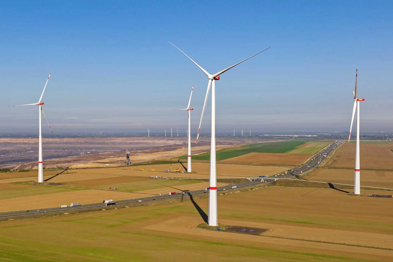 Luftaufnahme von Windkraftanlagen neben einer Autobahn, umgeben von Feldern unter einem klaren blauen Himmel.