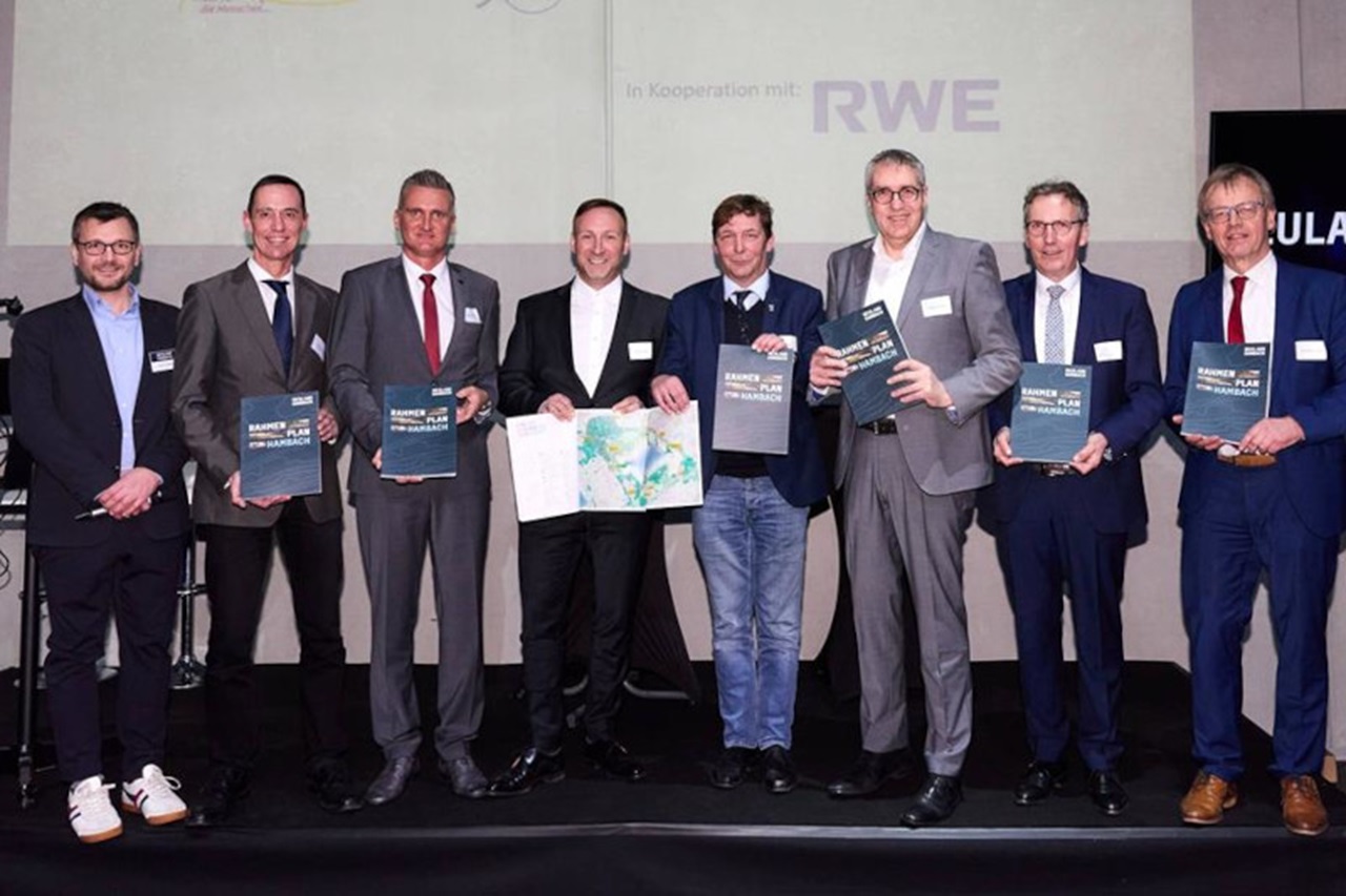 Eine Gruppe von acht Personen, die Bücher und eine Karte bei einer Veranstaltung halten. Sie stehen auf der Bühne vor einem neutralen Hintergrund.