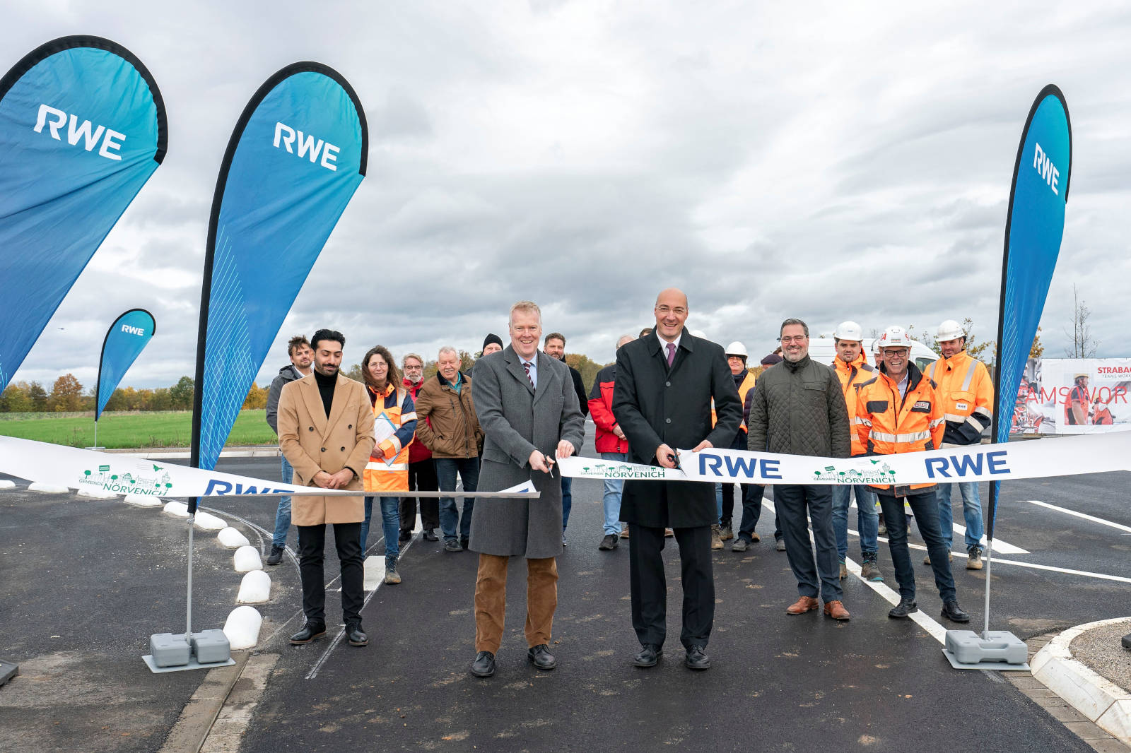 Zwei Männer schneiden ein RWE-Band durch, um eine neue Baustelle zu eröffnen. Im Hintergrund stehen Arbeiter in orangefarbener Kleidung.