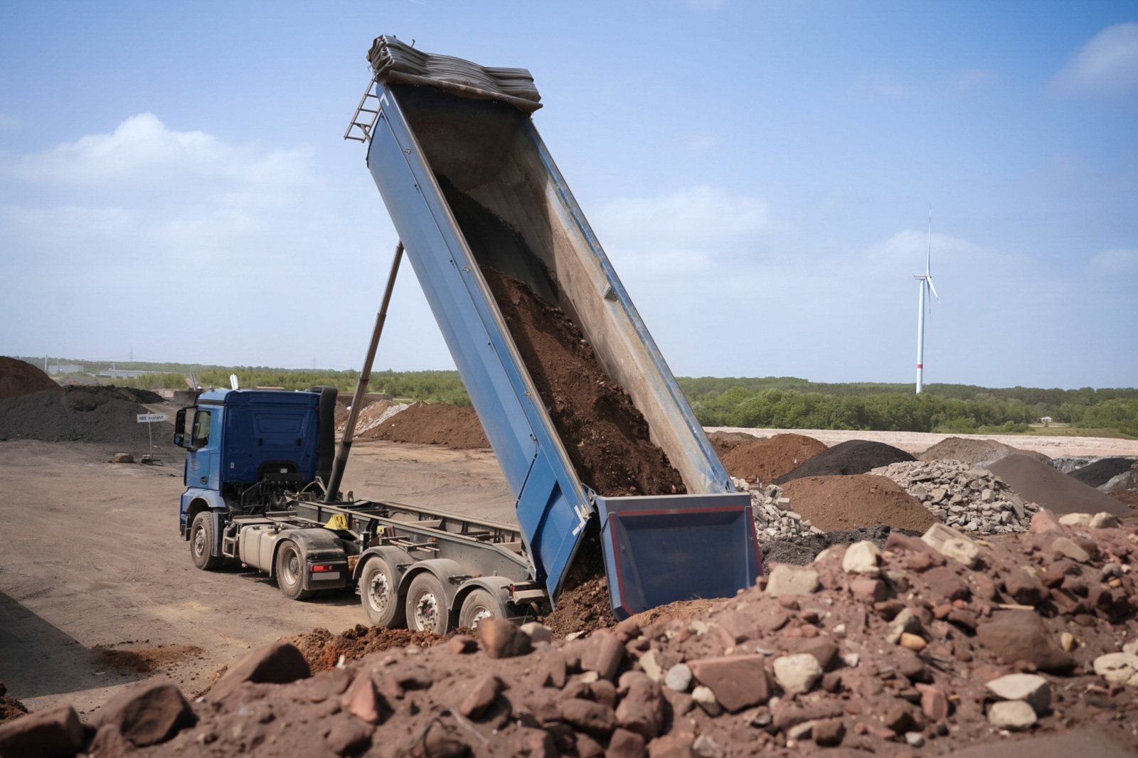 Ein blauer Lkw entlädt Erde auf einer Baustelle mit Erdhaufen und einem Windrad im Hintergrund.