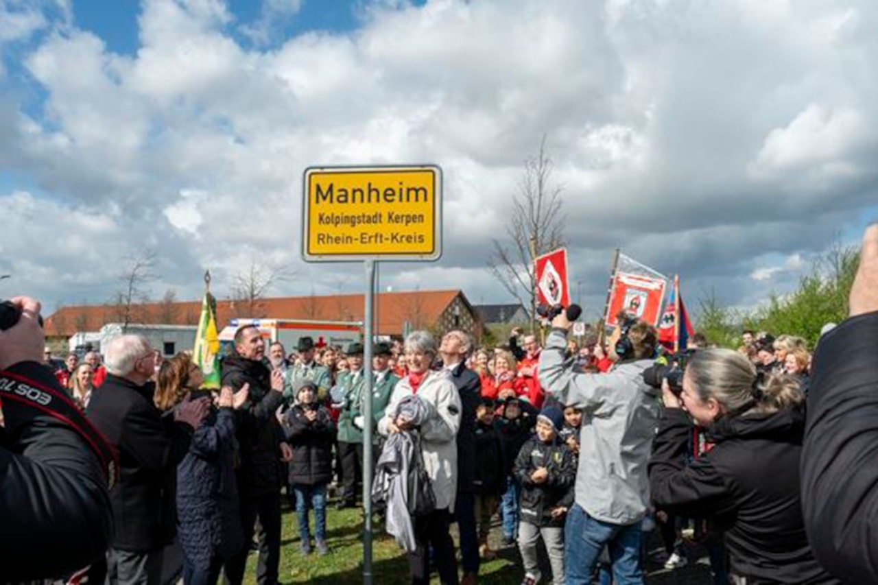 Eine Menschenmenge versammelt sich am Straßenschild für Manheim, feiert unter bewölkten Himmel mit Fahnen und Kameras.