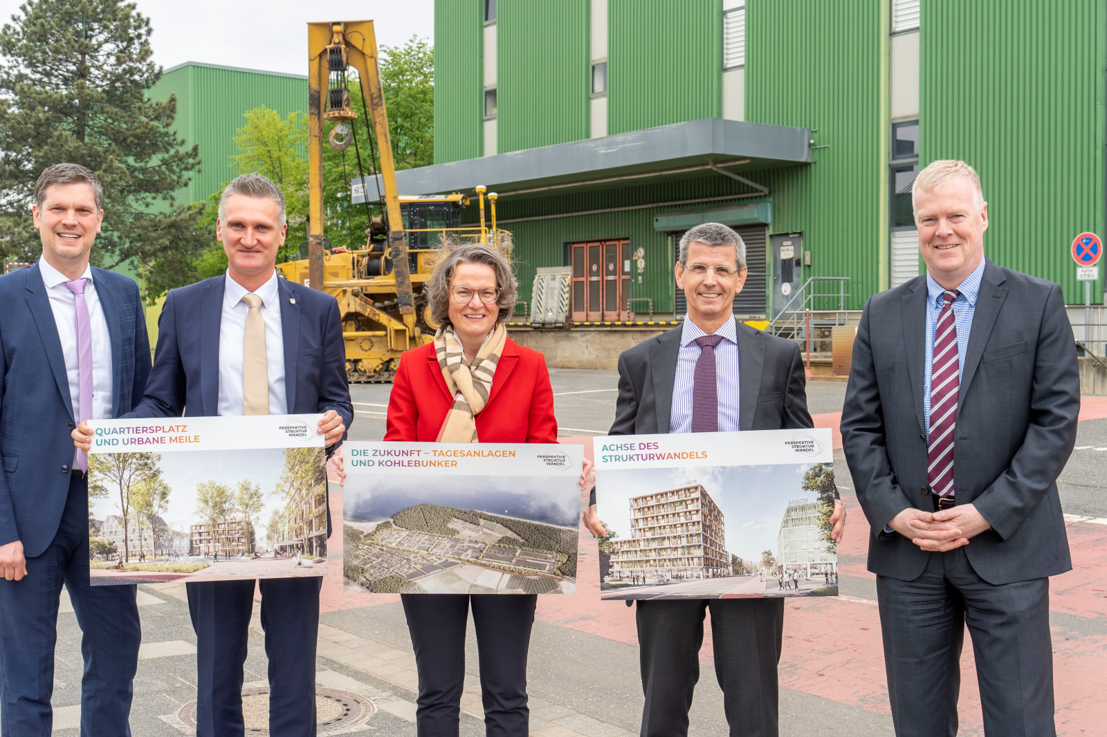 Gruppe von Fachleuten, die Projektpläne im Freien neben einem grünen Industriegebäude halten.
