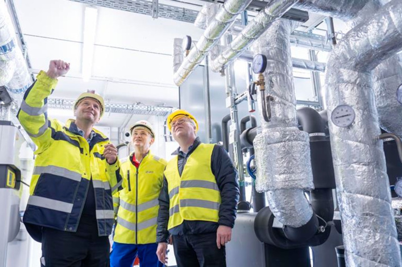 Drei Arbeiter in Sicherheitskleidung besprechen Geräte in einer technischen Einrichtung, umgeben von Rohren und Manometern.