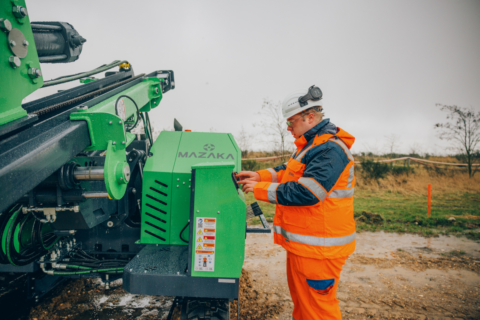 Ein Arbeiter in orangefarbener Sicherheitskleidung steht neben einer grünen Maschine und bedient diese unter bewölktem Himmel.