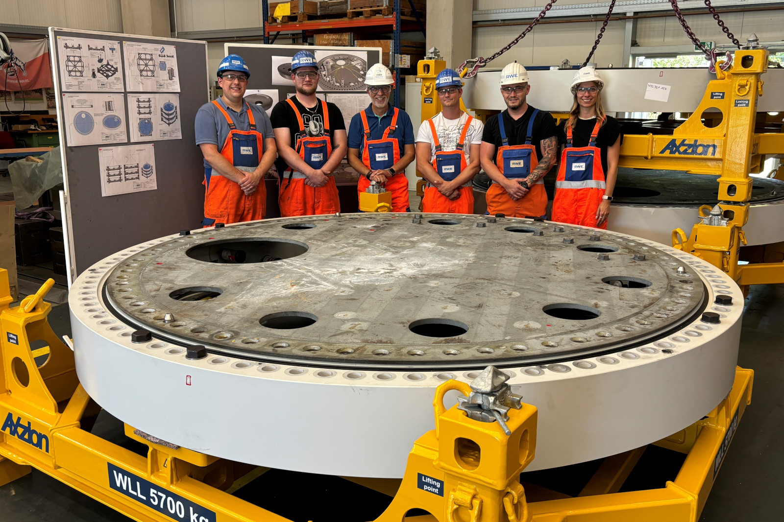 Eine Gruppe von Arbeitern in orangefarbenen Overalls steht neben einem großen kreisförmigen Industriebauteil in einer Werkstatt.