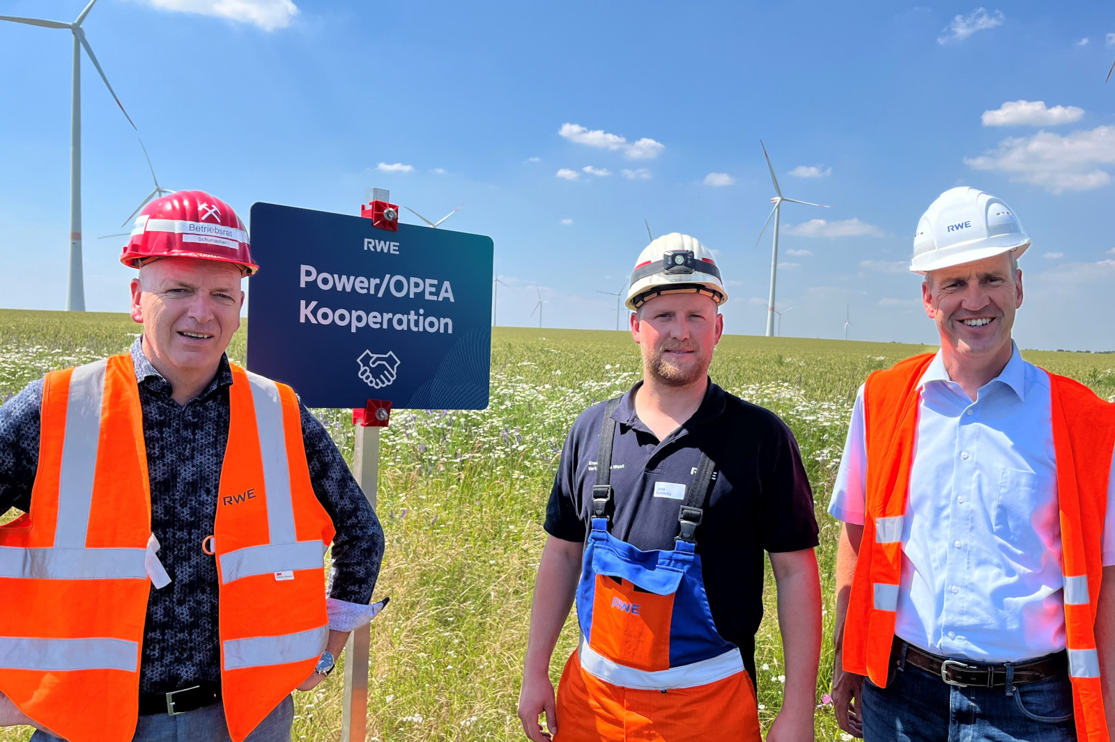 Drei Personen in Sicherheitskleidung stehen neben einem Schild zur RWE Power/OPEA Kooperation, mit Windturbinen im Hintergrund.