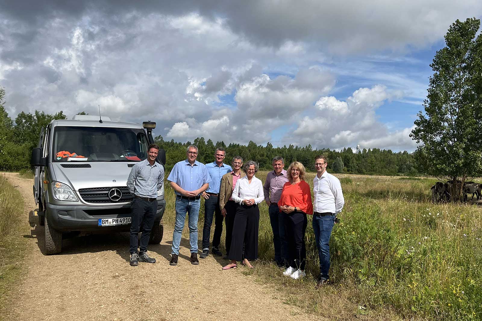 Eine Gruppe von acht Personen steht in der Nähe eines Mercedes-Vans auf einem Schotterweg, unter einem bewölkten Himmel mit Grünflächen.