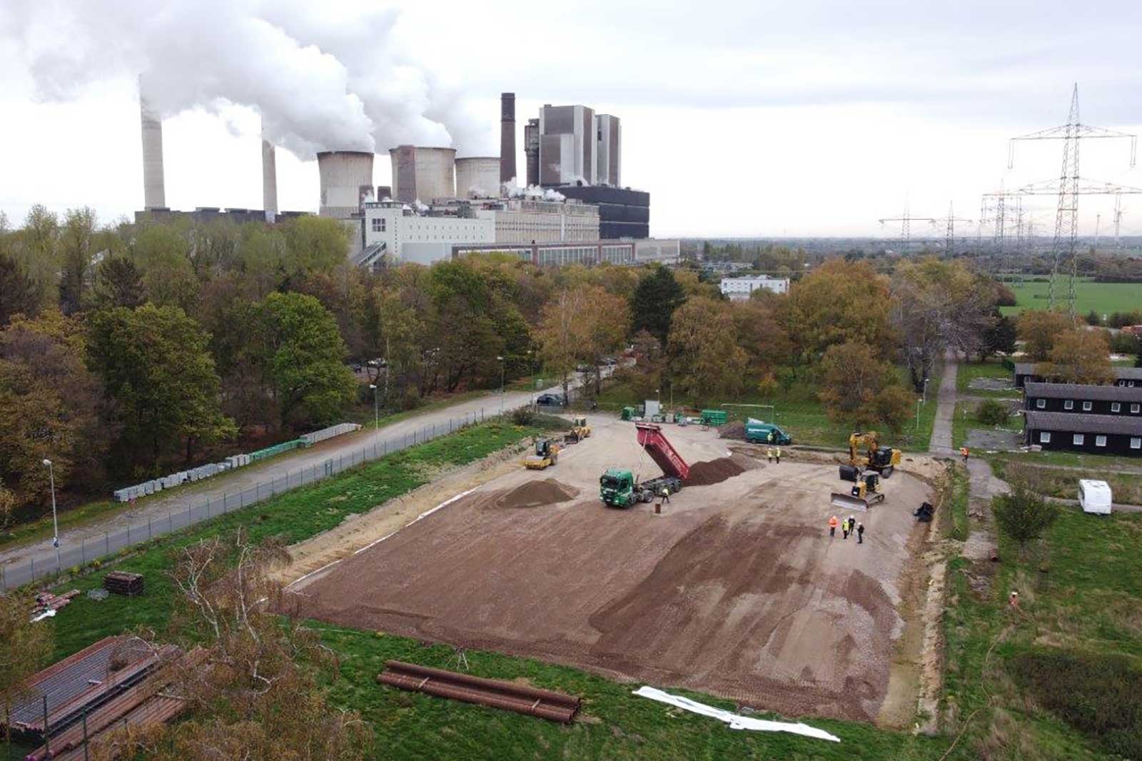 Eine Baustelle mit schwerem Gerät und Arbeitern, mit einem Kraftwerk und Schornsteinen im Hintergrund.