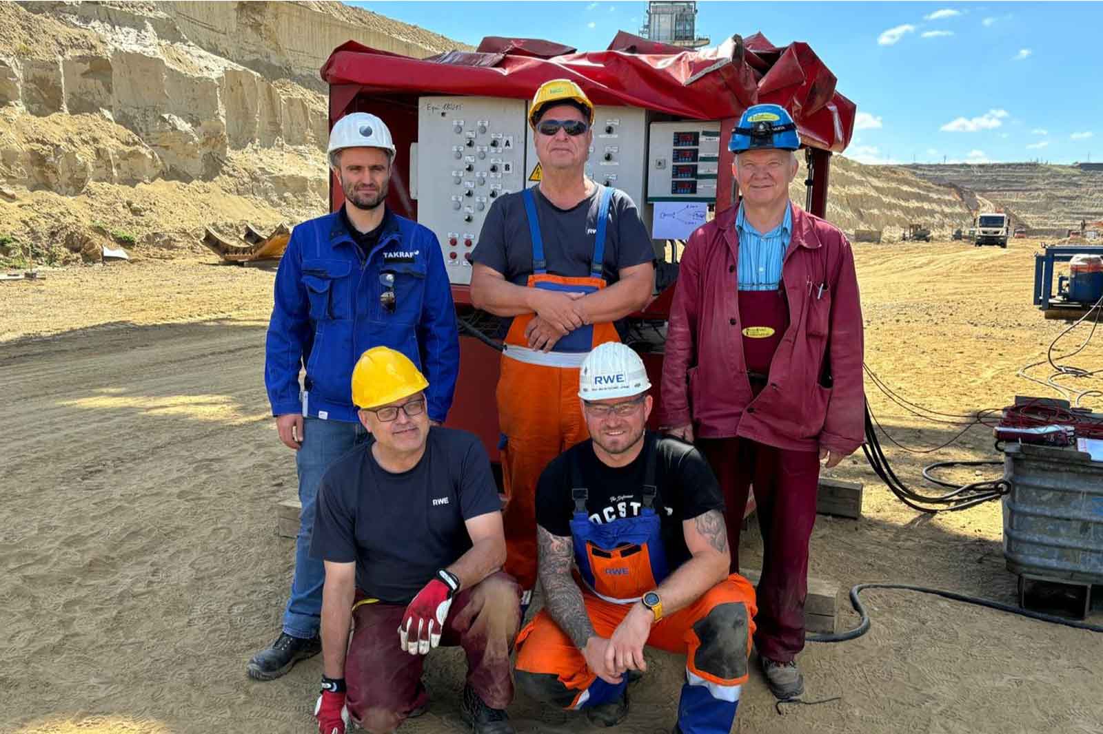 Fünf Arbeiter in Sicherheitskleidung stehen vor einer Maschine in einem Sandsteinbruch. Unter ihnen ist abgebauter Sand sichtbar.
