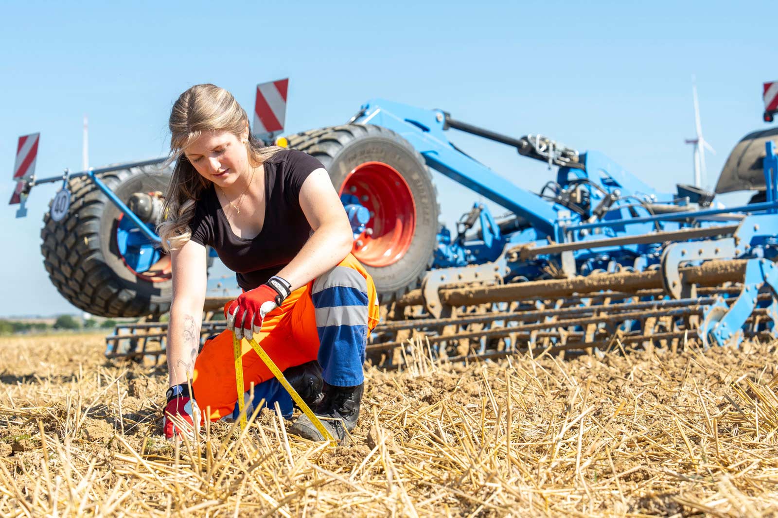 Eine Person kniet auf dem Boden und misst mit einem gelben Werkzeug neben landwirtschaftlichen Maschinen in einem sonnigen Feld.