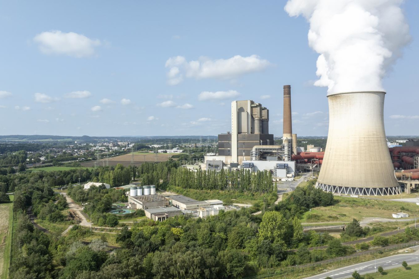 Ein Industriekraftwerk mit einem Kühlturm, der Dampf abgibt, umgeben von Grünflächen und Feldern unter einem blauen Himmel.