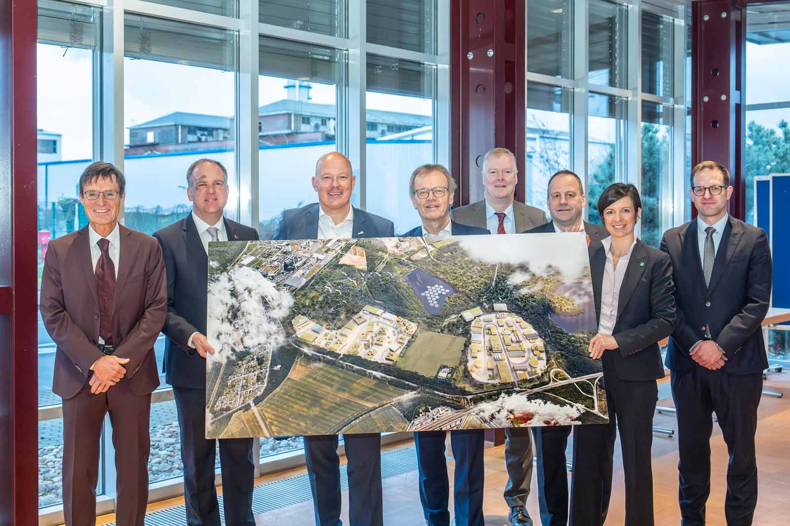 Eine Gruppe von Personen hält eine Luftbildkarte eines Entwicklungsstandorts mit Gebäuden und Landschaften.