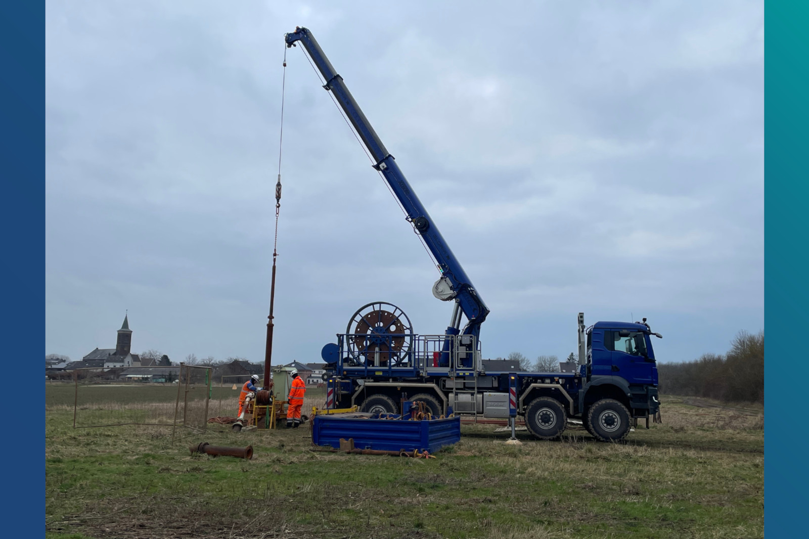 Ein blauer Kran hebt Ausrüstung an, während zwei Arbeiter in orangenen Sicherheitsanzügen auf einem Feld in der Nähe eines Kirchturms stehen.