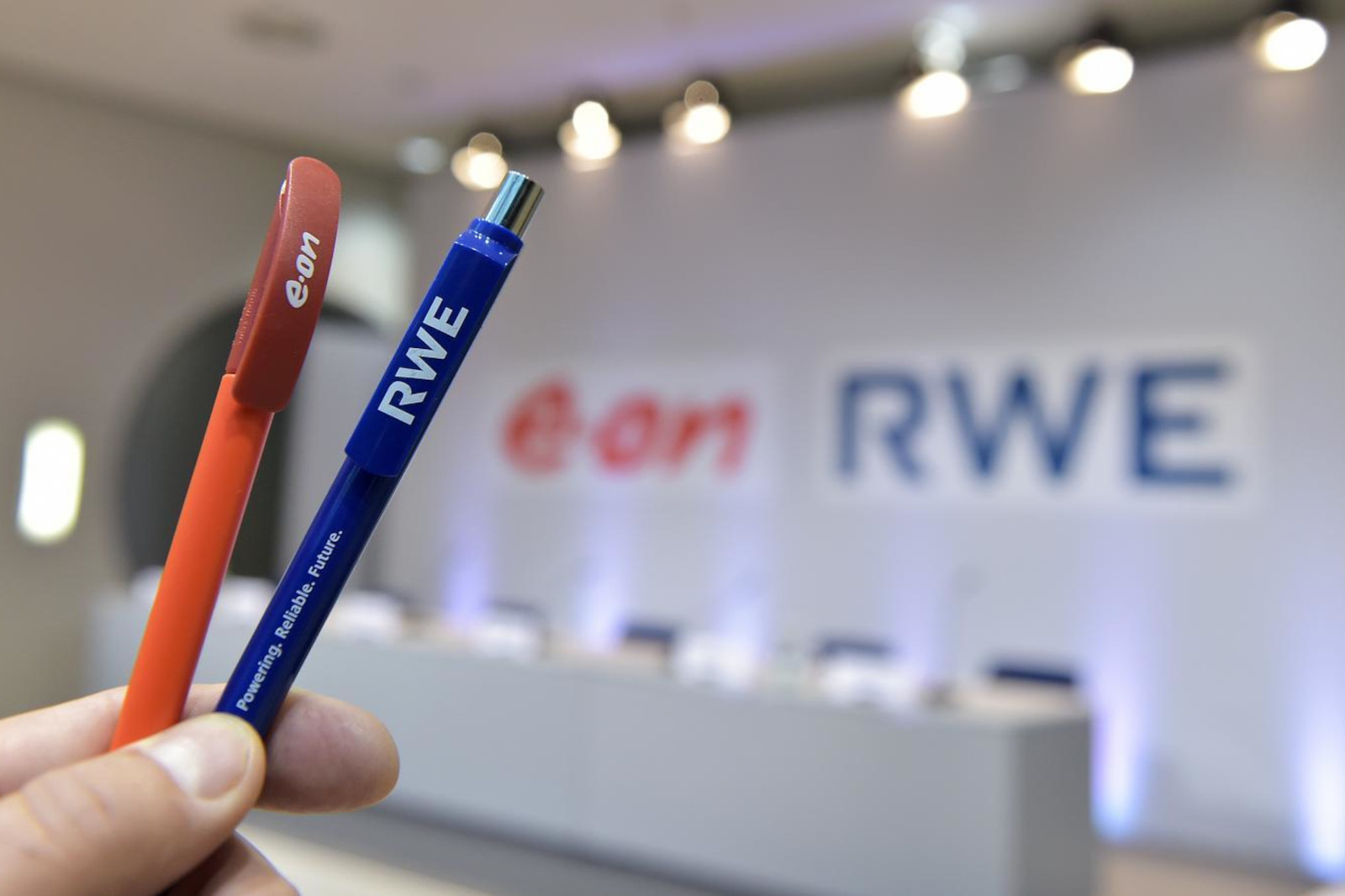 Eine Person hält einen roten e.on Stift und einen blauen RWE Stift vor einem Konferenzhintergrund mit beiden Firmenlogos.
