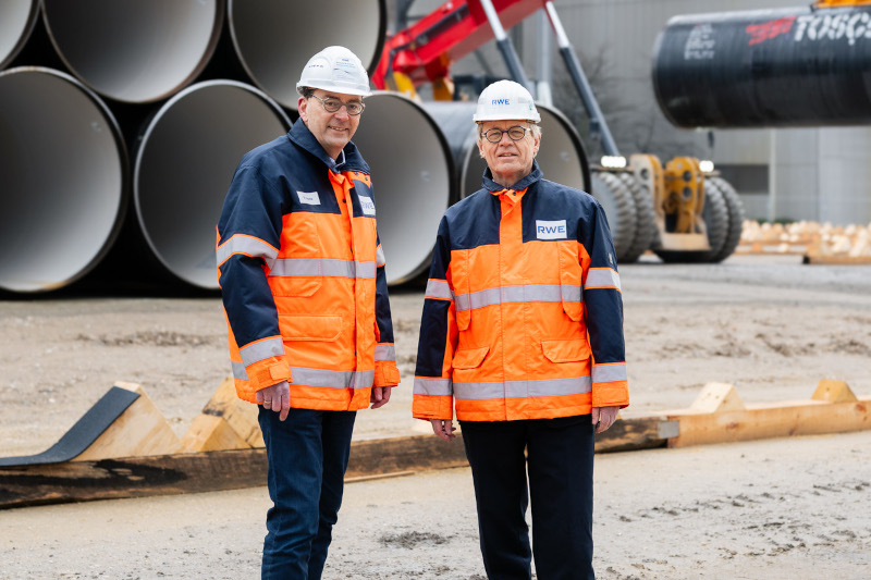 Zwei Arbeiter in hochsichtbaren Jacken stehen vor großen Rohren auf einer Baustelle.
