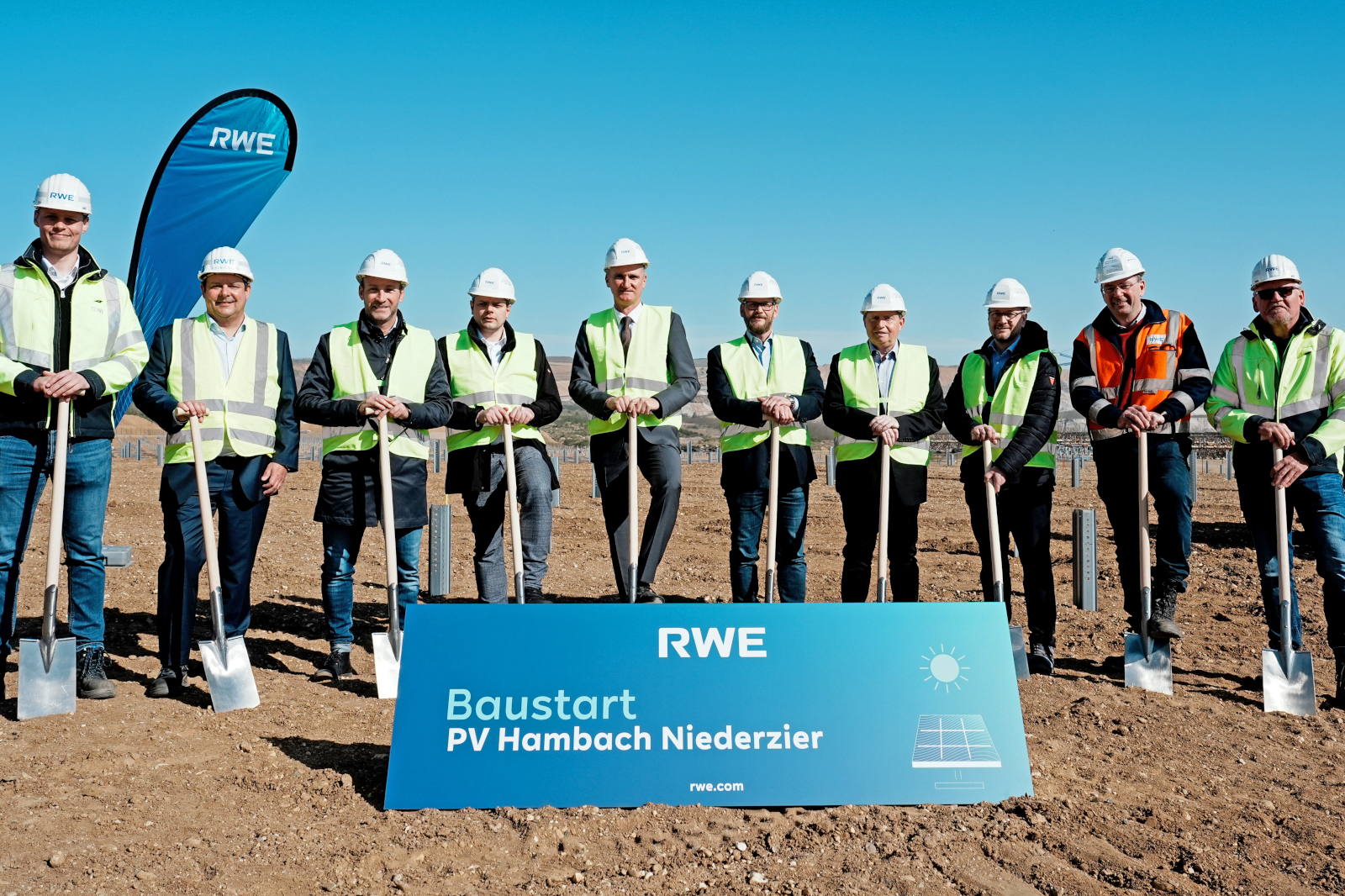 Eine Gruppe von Personen in Sicherheitswesten und Helmen bei einer Grundsteinlegung für ein Solarprojekt, mit einem blauen Schild, das Projektdetails anzeigt.