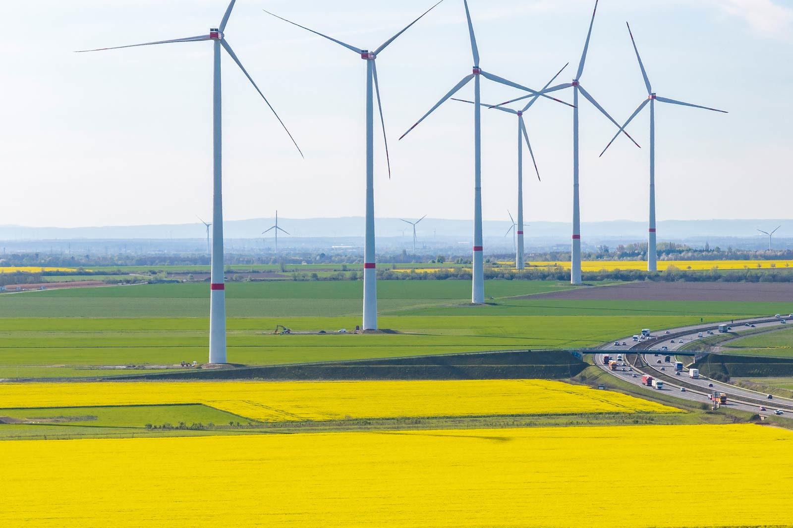 Windkraftanlagen ragen über lebhaften gelben Feldern, mit einer kurvenreichen Straße und Fahrzeugen im Vordergrund.