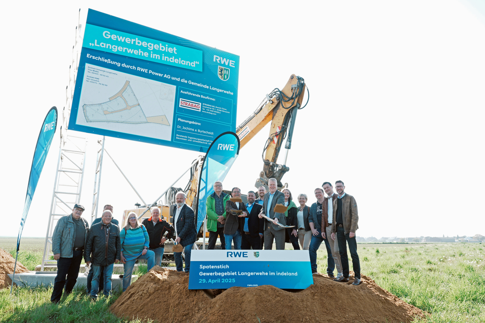 Eine Gruppe von Menschen bei einer Grundsteinlegung mit einem großen Bagger und einem Schild, das die Projektdetails in Langerwehe anzeigt.