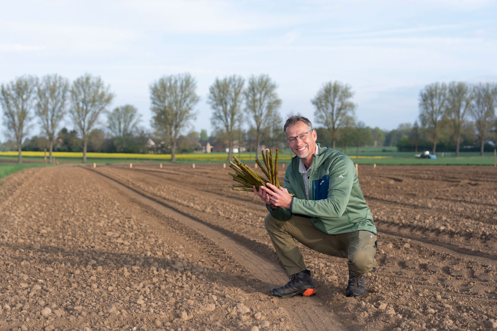 Ein Landwirt hockt auf dem bearbeiteten Boden und hält frisch geerntete Spargelstangen in einem Feld mit Bäumen im Hintergrund.