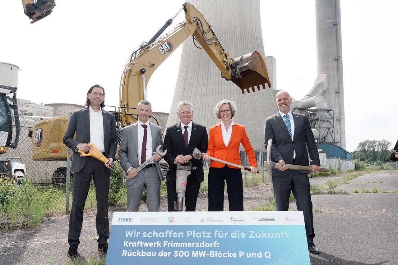 Eine Gruppe von fünf Personen, die Bauwerkzeuge hält, steht vor einem Kraftwerk mit Maschinen und einem Schild.