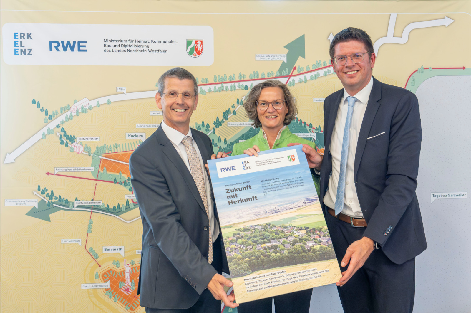 Drei Personen halten ein Plakat mit dem Titel 'Zukunft mit Herkunft' vor einer Karte, die ein Gemeinschaftsentwicklungsprojekt darstellt.