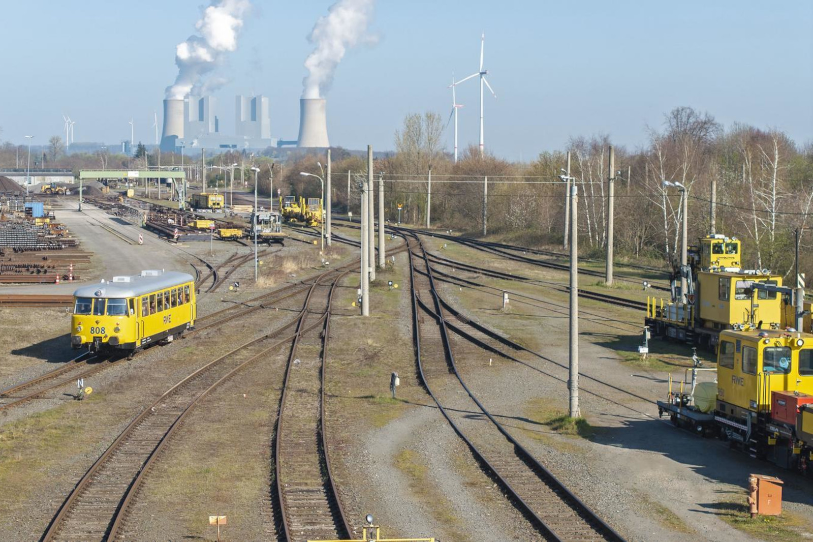 Ein gelber Zug auf Gleisen neben Industriegebäuden und Windkraftanlagen, mit Rauch, der aus Kühltürmen aufsteigt.