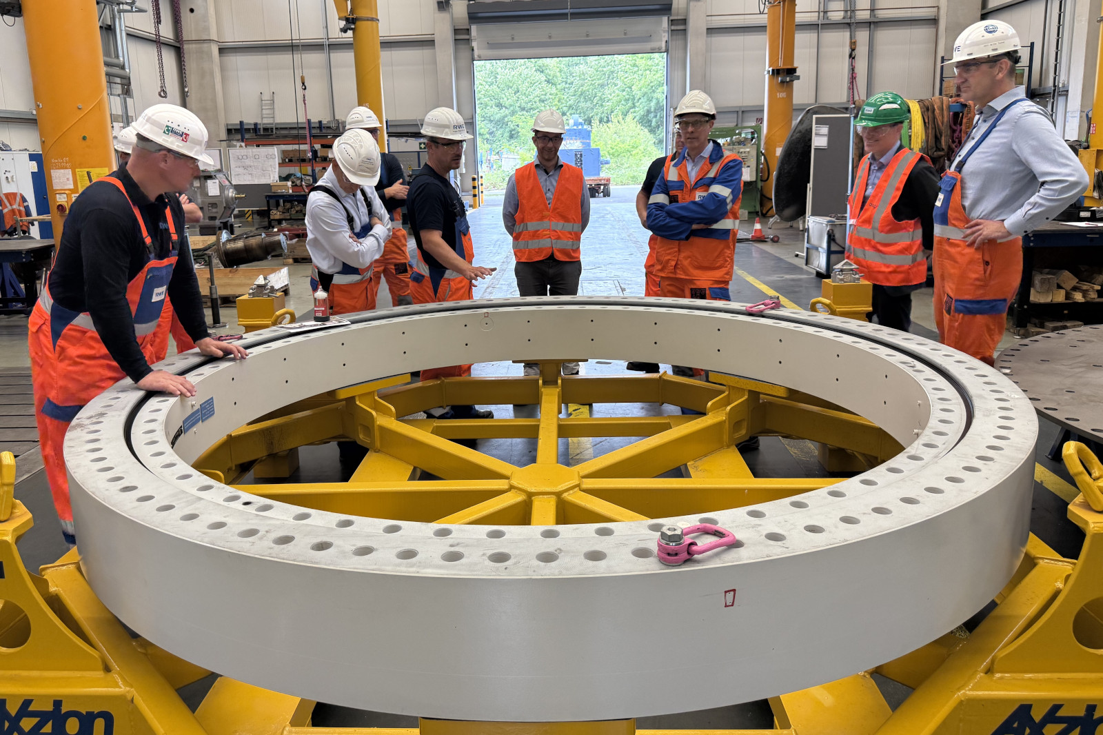 Eine Gruppe von Arbeitern in Sicherheitskleidung beobachtet ein großes kreisförmiges Bauteil auf einer gelben Maschine in einer Werkstatt.