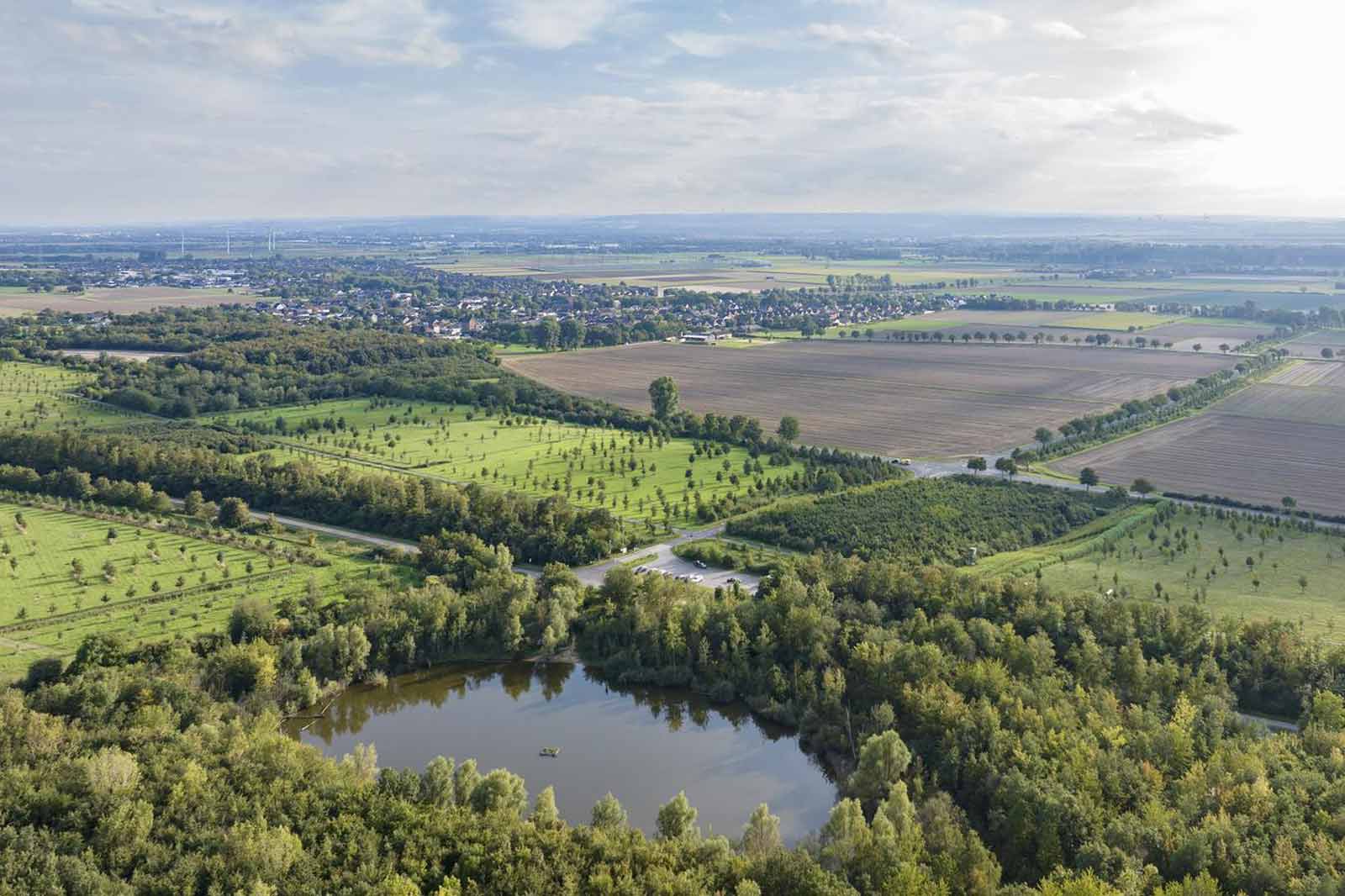 Luftaufnahme von grünen Feldern, einem kleinen See und einem Dorf in der Ferne unter einem bewölkten Himmel.