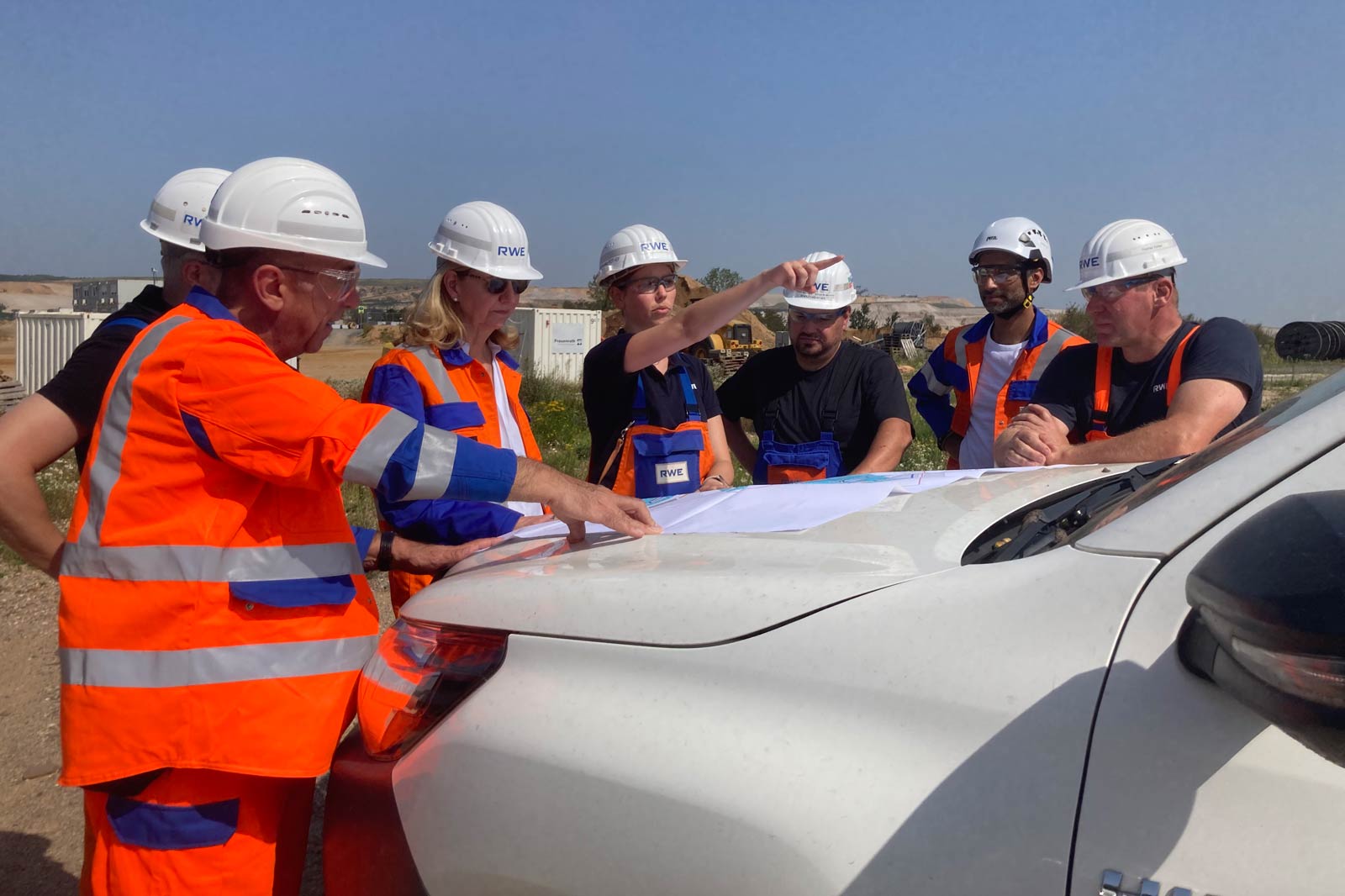 Eine Gruppe von Fachleuten diskutiert Pläne über eine Karte auf einem Fahrzeug in einem sonnigen Freien.