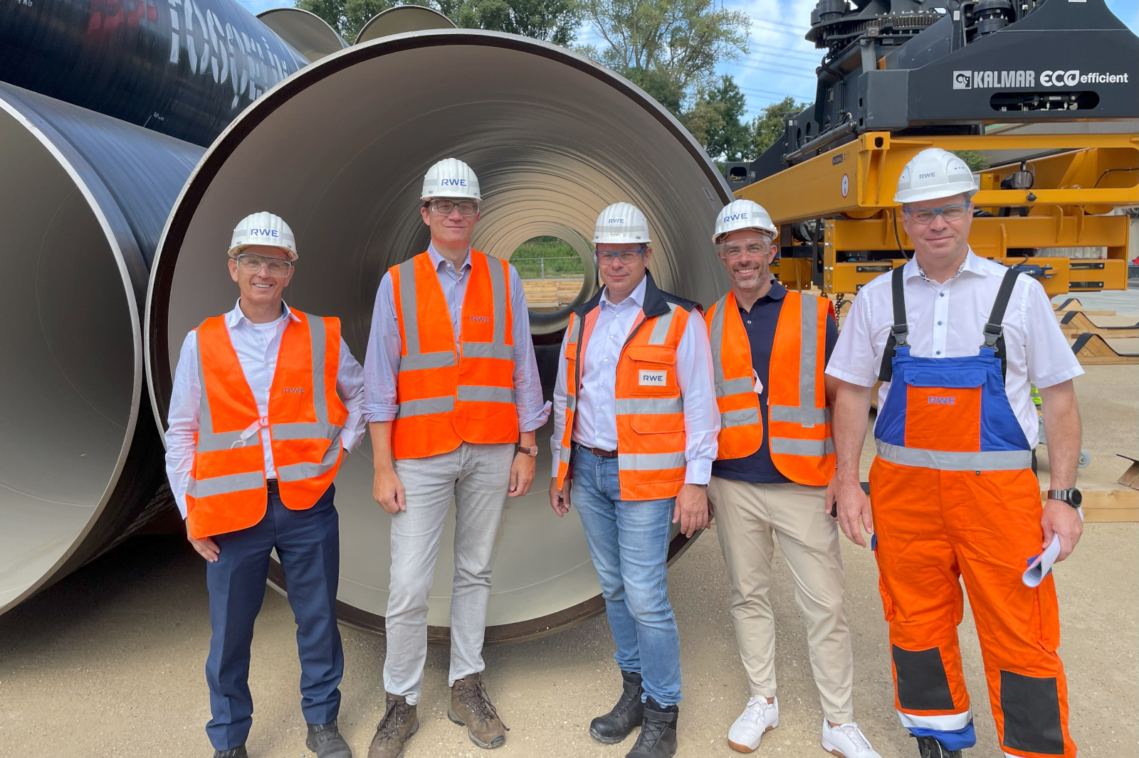 Fünf Personen in Sicherheitskleidung stehen zusammen, mit großen Rohren und Maschinen im Hintergrund.