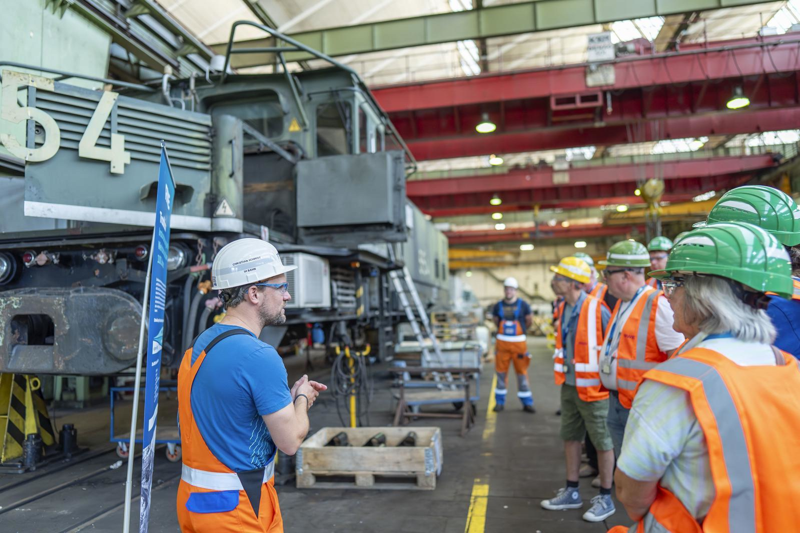 Eine Werkstatt-Szene mit Mitarbeitern in Schutzausrüstung. Der Fokus liegt auf einer Diskussion über Maschinen, einschließlich eines großen Fahrzeugs.