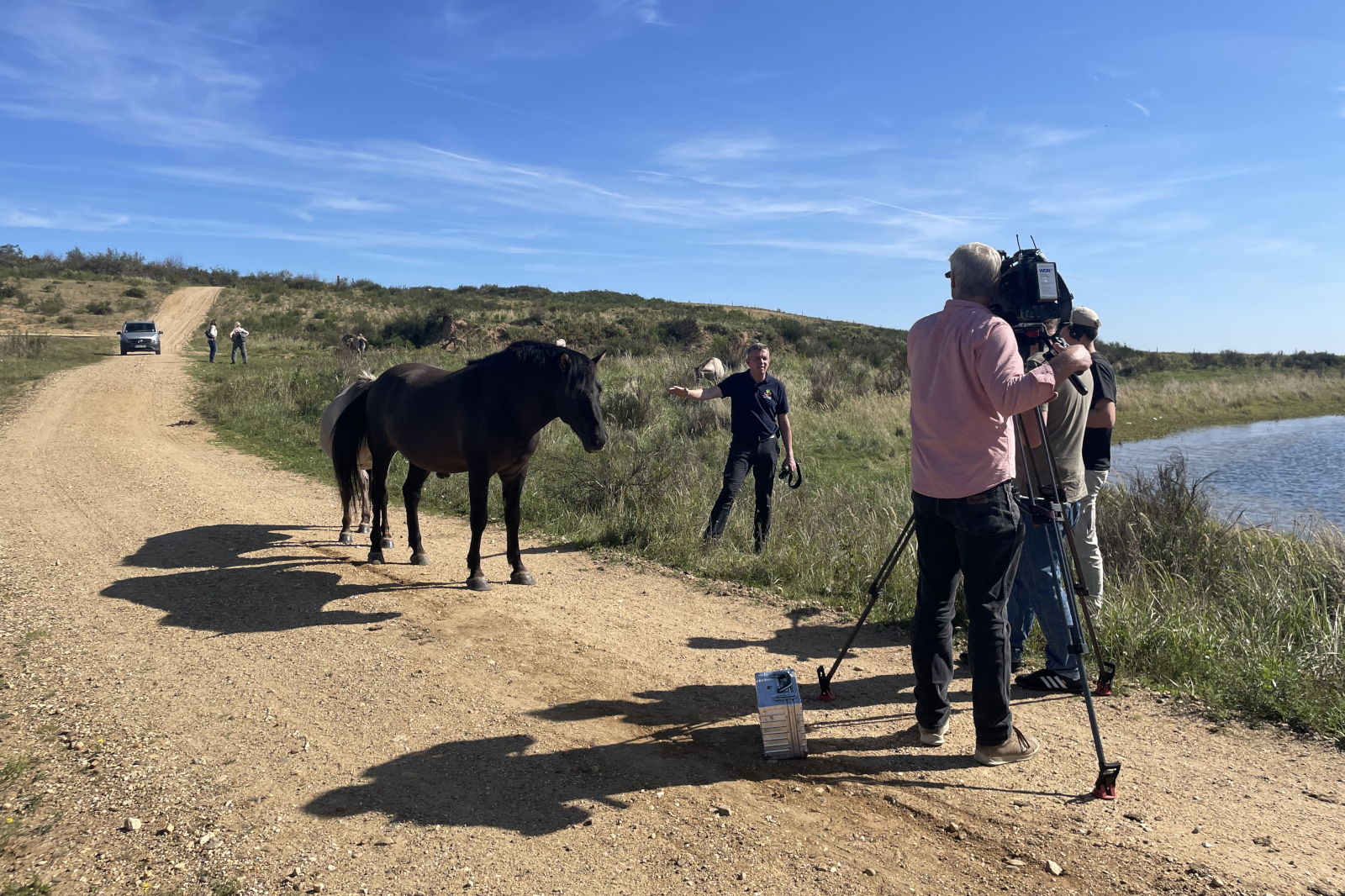 Ein Filmteam filmt eine Szene mit zwei Pferden in der Nähe eines Gewässer und einer Schotterstraße unter einem klaren blauen Himmel.