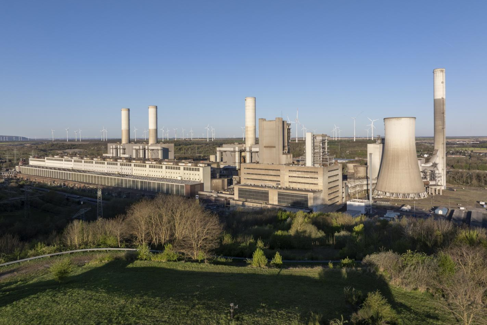 Ein Industriekomplex mit Kühltürmen und Schornsteinen, gelegen in der Nähe von Windkraftanlagen unter einem klaren blauen Himmel.
