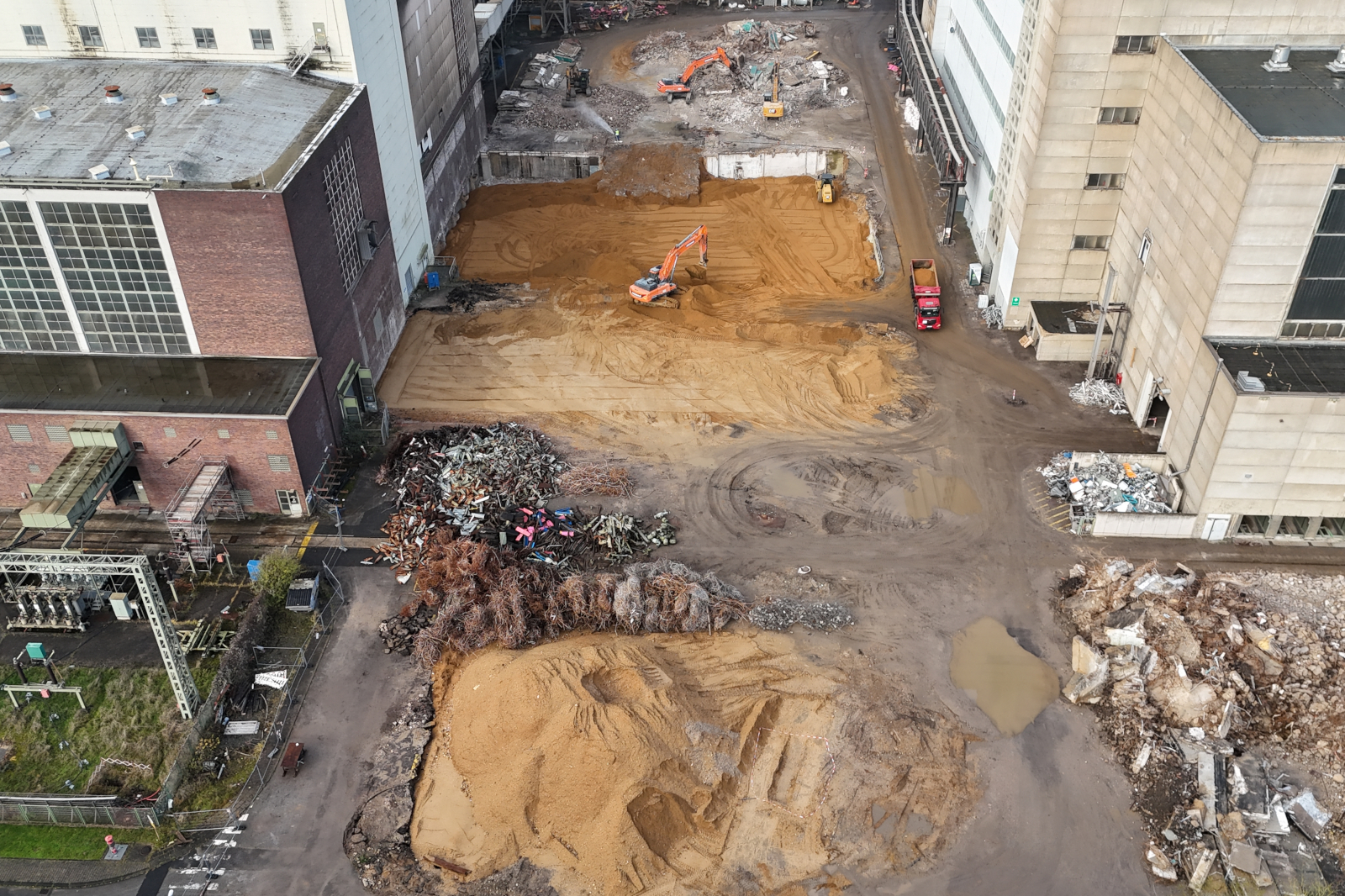 Eine Luftaufnahme einer Baustelle mit Maschinen, die Erde bewegen und Haufen von Schrott. Sand bedeckt den Boden, es gibt Pfützen.