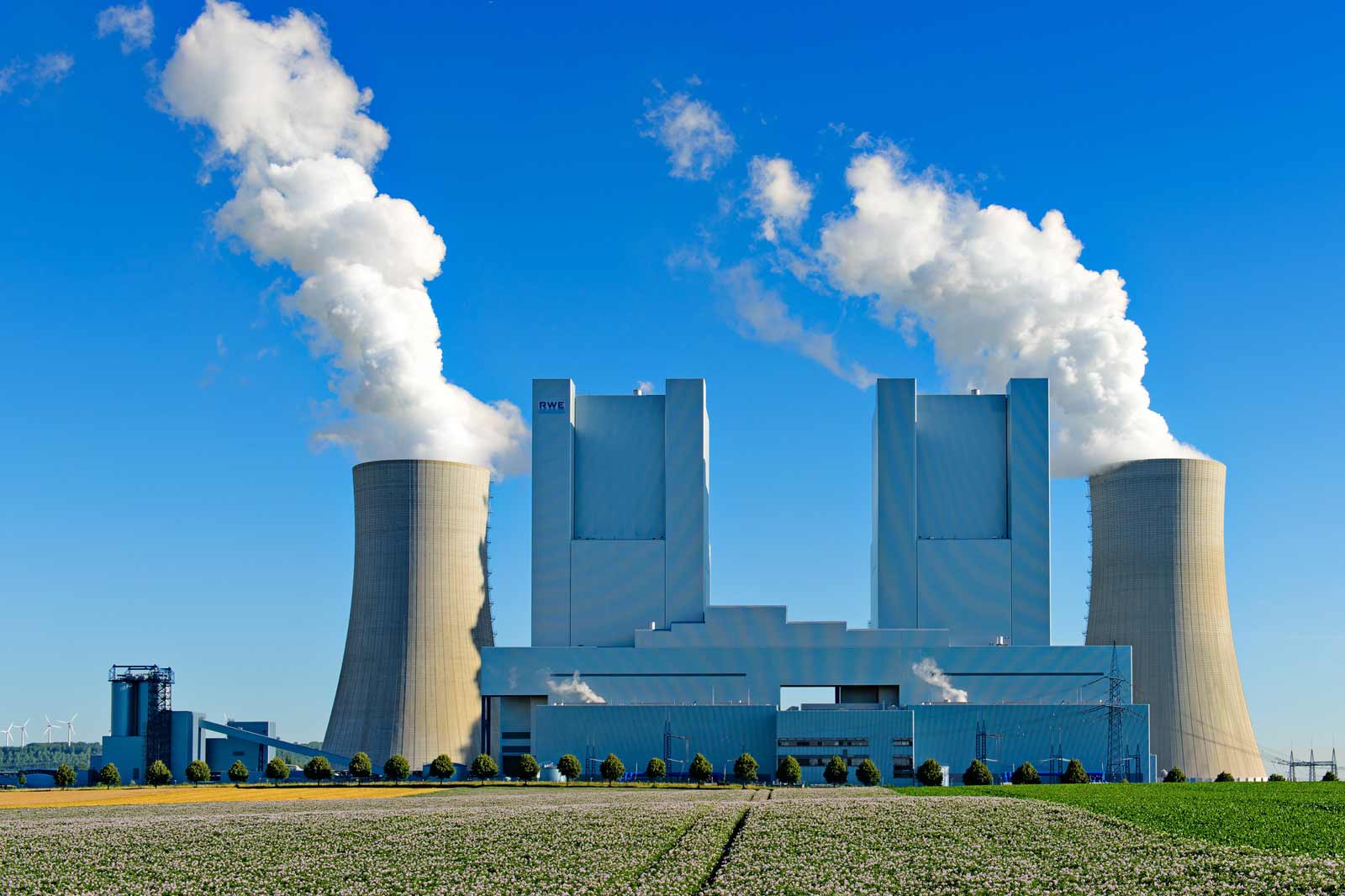 Ein großes Kraftwerk mit Kühltürmen, die Dampf gegen einen klaren blauen Himmel ausstoßen, umgeben von grünen Feldern.