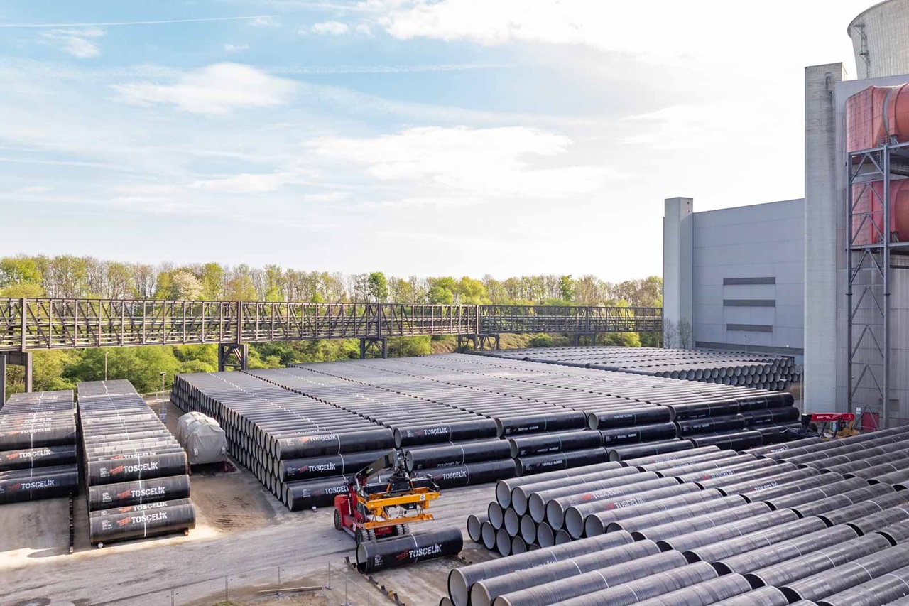 Luftaufnahme einer Baustelle mit gestapelten schwarzen Rohren und einem großen Industriegebäude im Hintergrund.