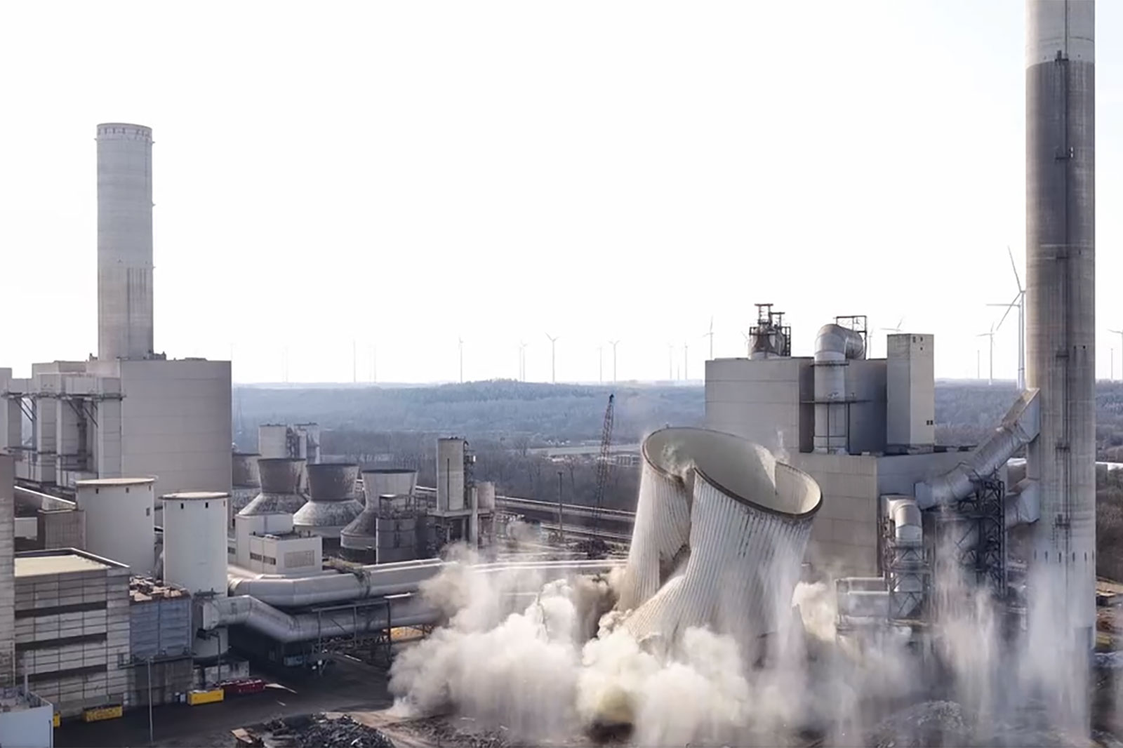 Zwei eingestürzte Kühltürme eines Kraftwerks erzeugen eine große Staubwolke, umgeben von Gebäuden, hohen Schornsteinen und Windrädern am Horizont.